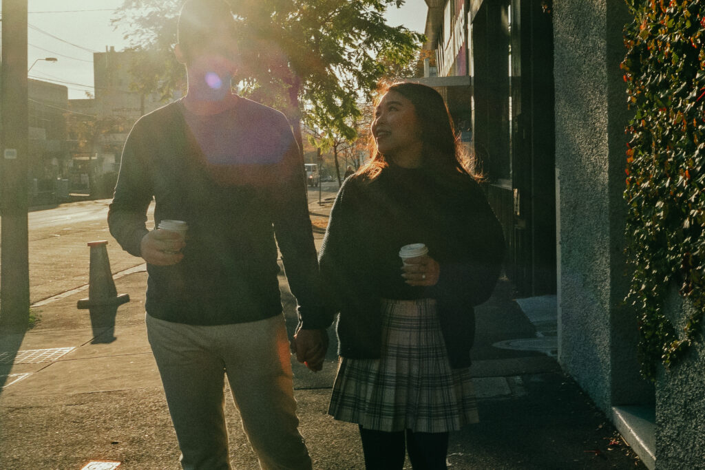 couple holding hands and talking while holding coffees, the sun rays are shining behind them