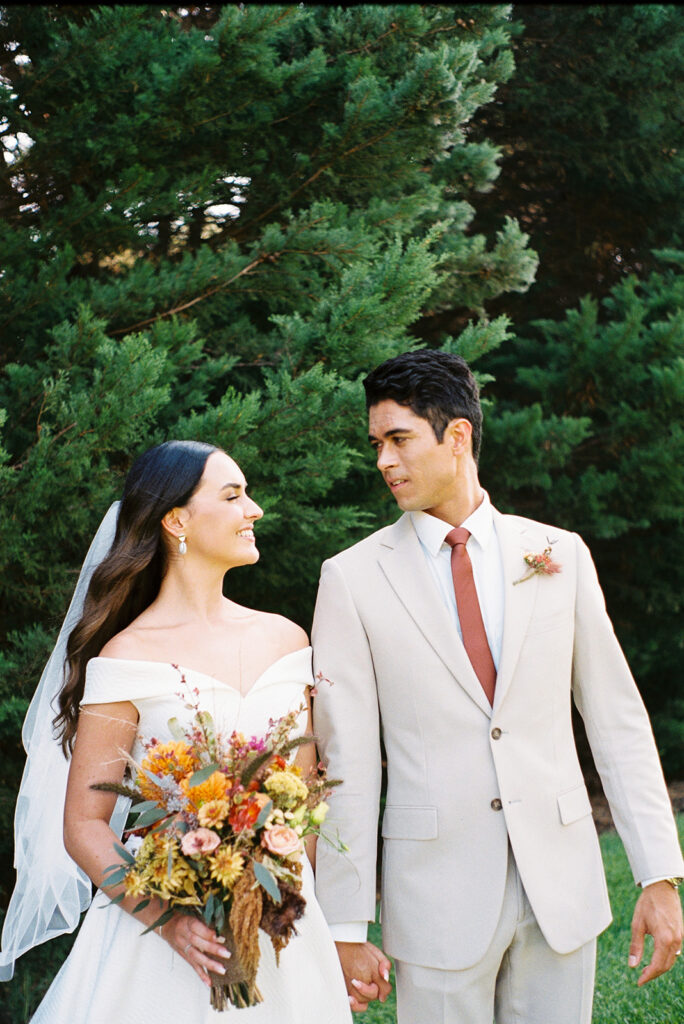 Bride and groom staring into each other's eyes during their wedding portraits in front of a large bush and lush green lawn captured on film