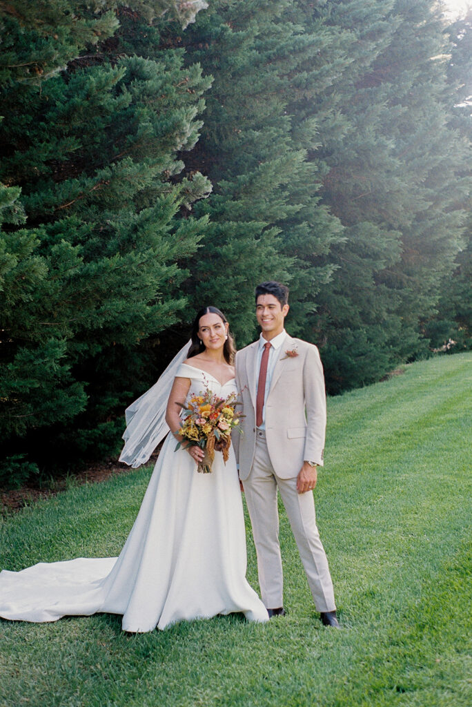 Bride and groom smiling together while holding hands during their wedding portraits in front of a large bush and lush green lawn captured on film