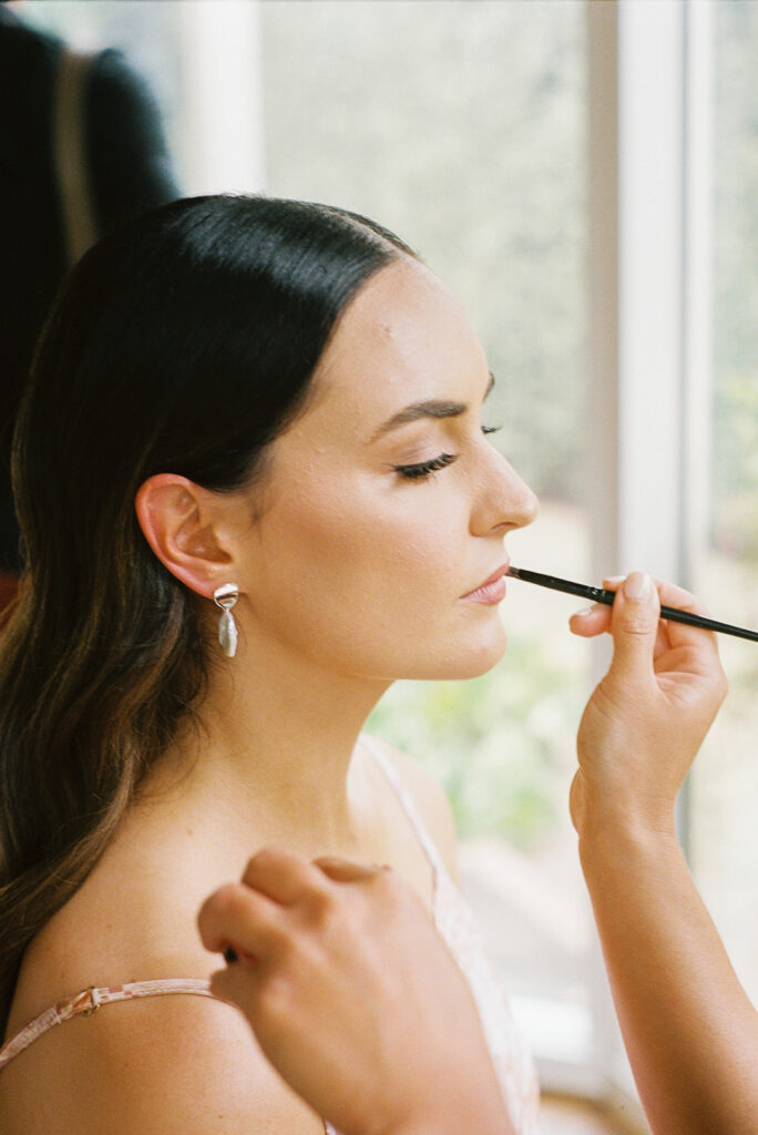 Bride is getting her lipstick touched up while sitting in front of a large window