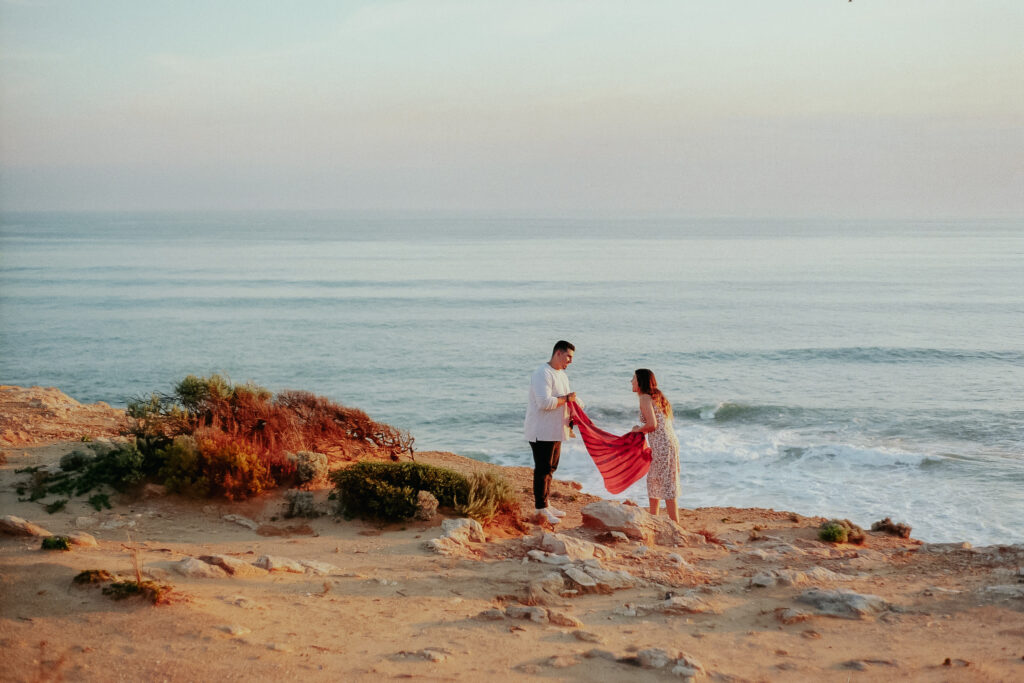 Couple laying out a picnic blanket together on top of a cliff in front of beach views during their engagement session 