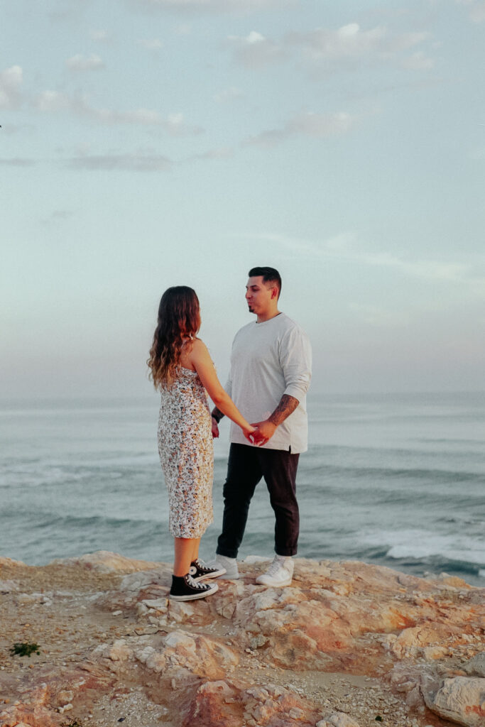 Couple holding hands on top of a cliff in front of beach views during their engagement session captured on film