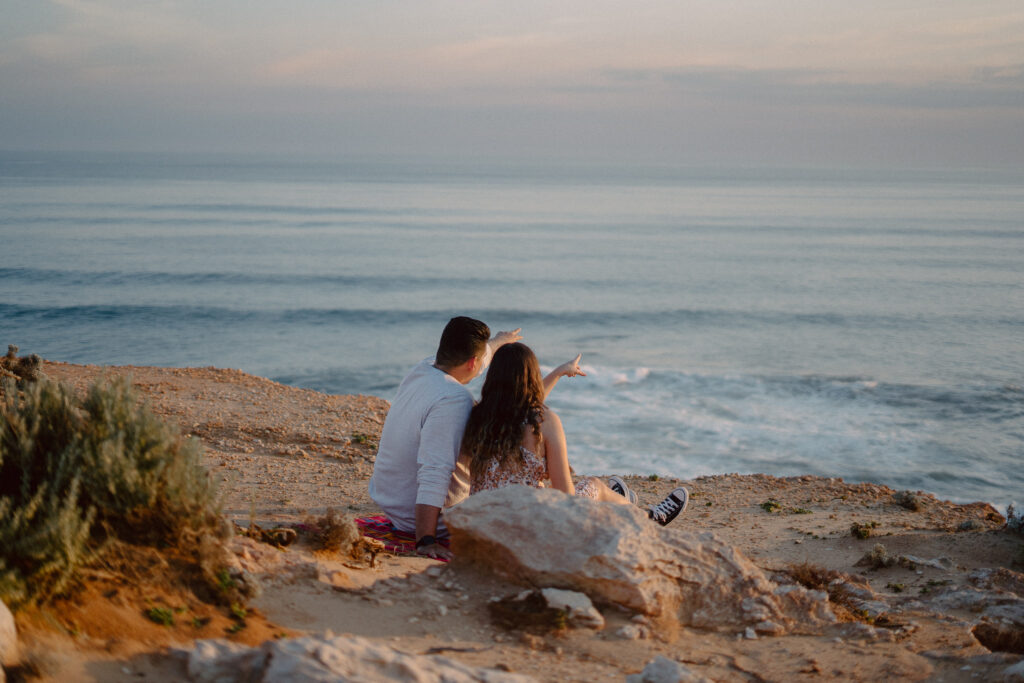 Couple pointing out into the ocean together, sitting on a picnic blanket together on top of a cliff in front of beach views during their engagement session 