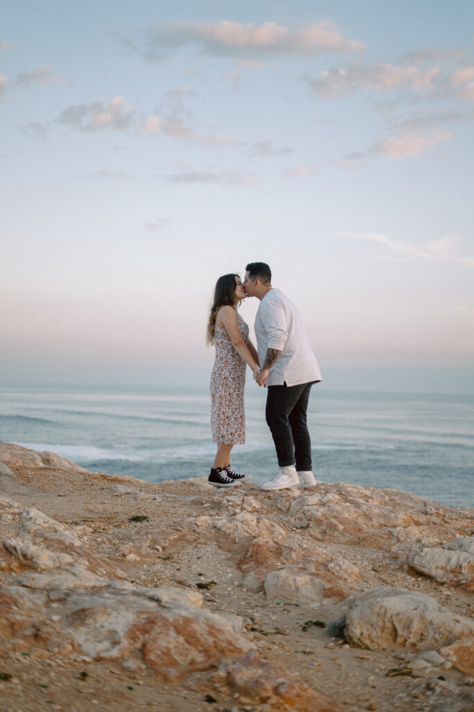 Couple kissing on top of a cliff in front of beach views during their engagement session 