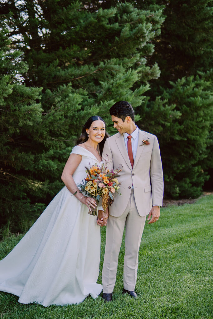 Bride and groom smiling at each other during their wedding portraits in front of a large bush and lush green lawn