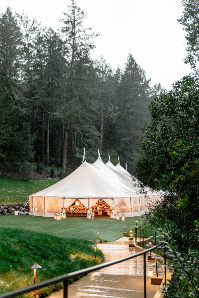 A white marquee glowing with light in the middle of a forest with a leading pathway for guest to walk 