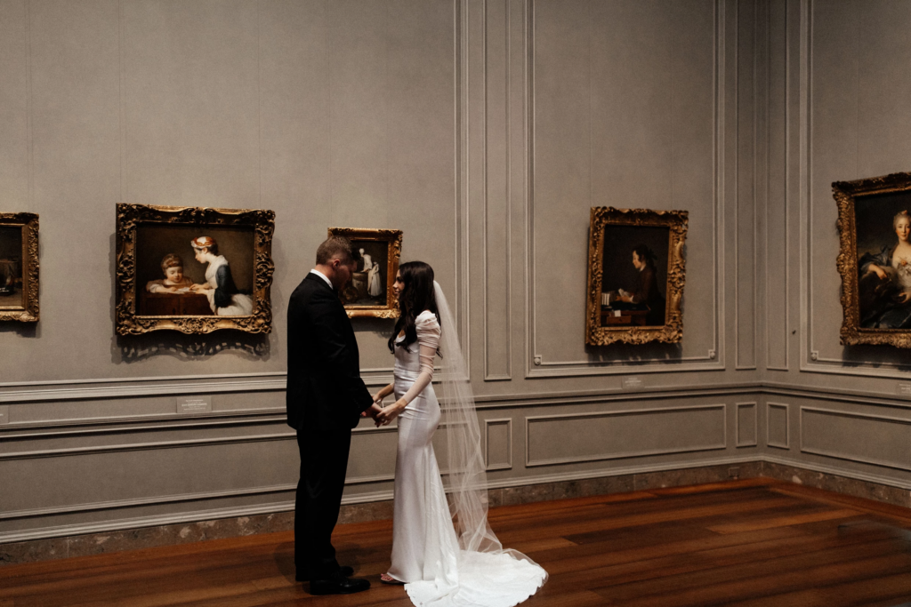 A bride and groom holding hands in an art gallery