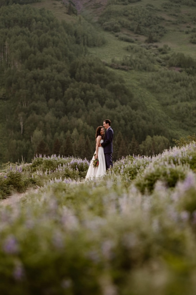 A bride and groom surrounded by purple flowers with mountain view behind them