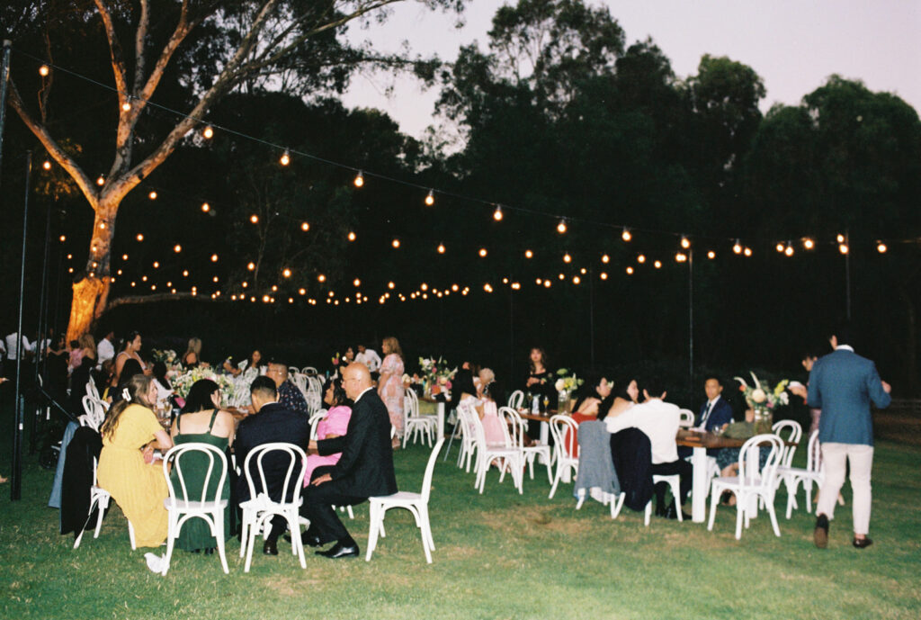 Guests seated outside under the festoon lights during a wedding reception at emu bottom homestead, captured on 35mm film