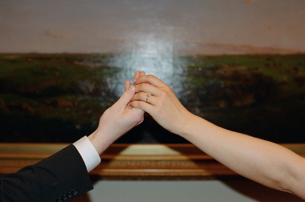 35mm photo of a couple holding hands in front of a painting, an engagement ring is visible