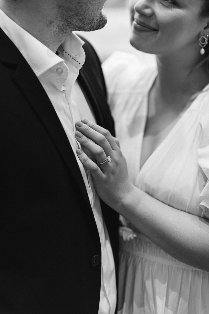 Close up of a couple smiling at each other, an engagement ring is in focus where her hand is laying on his chest