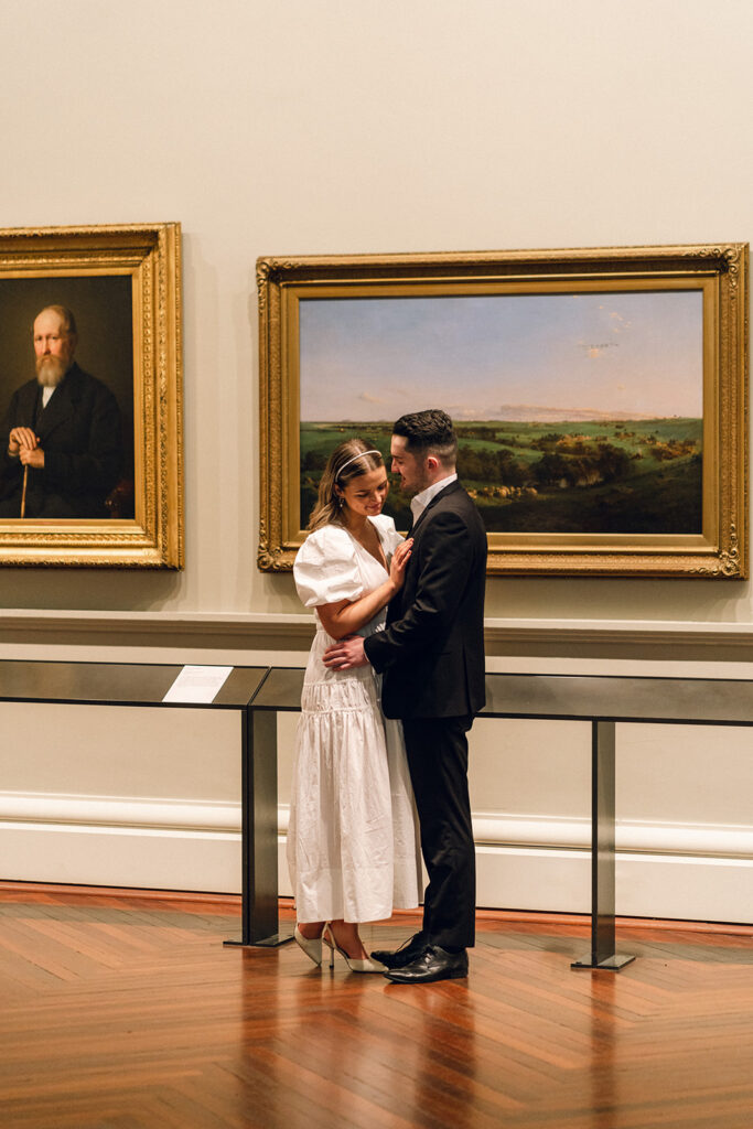 Couple is cuddling, standing in front of two paintings in the melbourne state library gallery, she is wearing a long white dress and he is wearing a black suit.