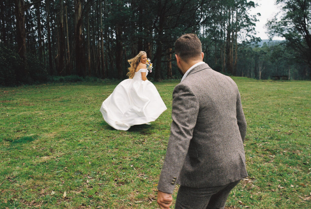 a bride playfully running away from a groom during their wedding portraits session at toolangi heights, the wind is blowing through her long blond hair and her white silky wedding dress