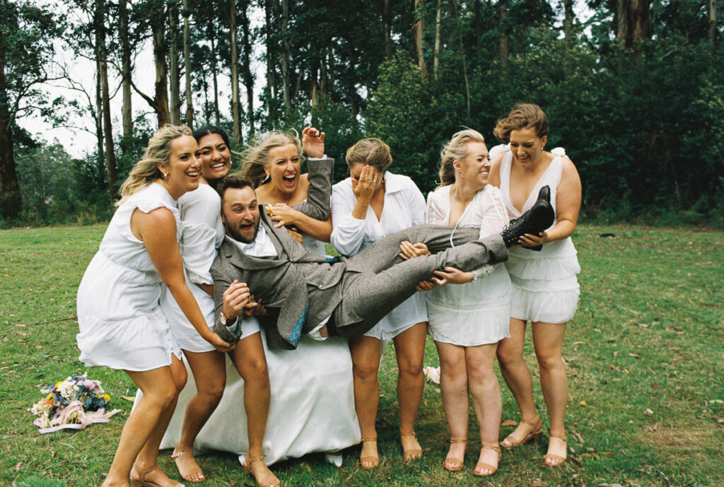 a film photo of a bride and her bridesmaids lifting up the groom, wind is blowing in their faces and they are smiling and laughing