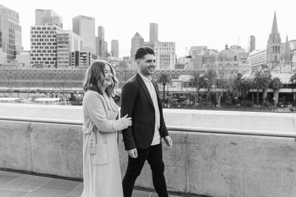 a black and white photo of a couple holding hands during their pre-wedding photos, walking and laughing together with the southbank melbourne cityscape behind them