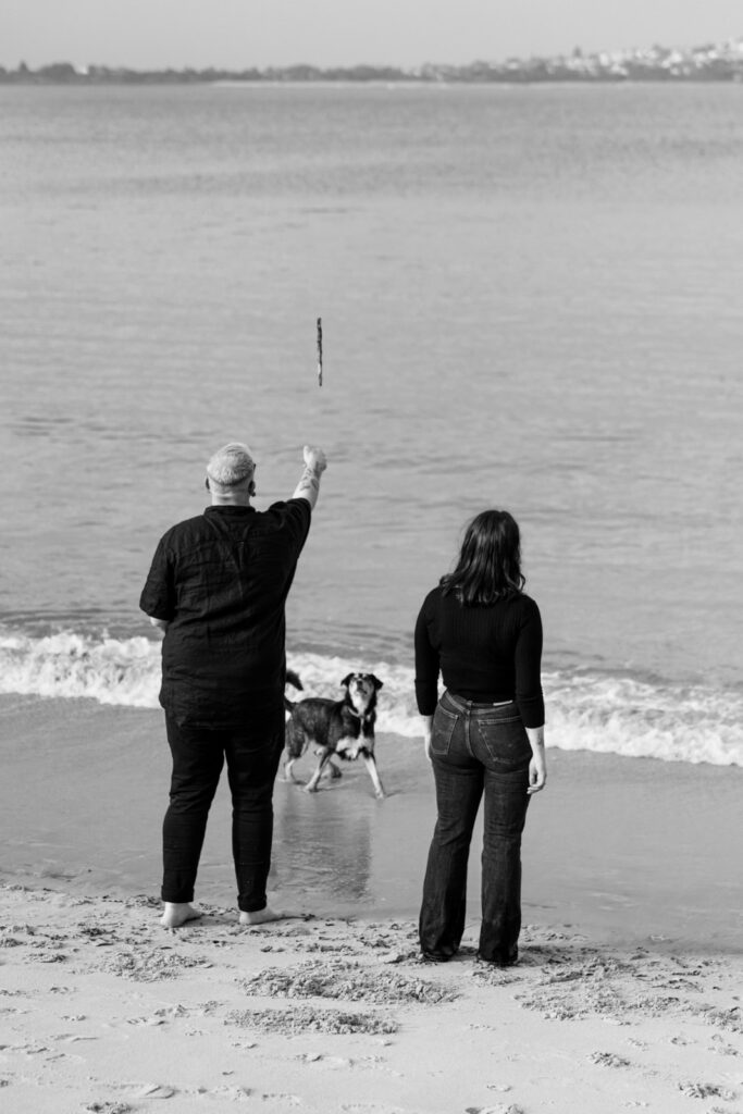 a black and white photo of a couple throwing a stick in the air for their dog to catch in the water on the shore of a beach