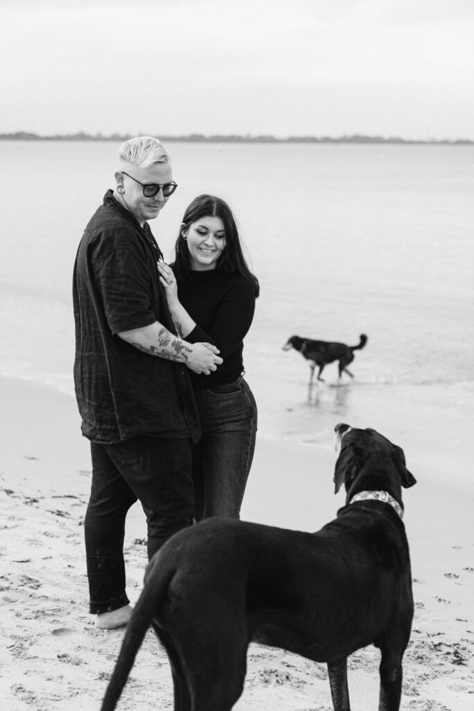 couple hugging during their engagement photos and staring at their dog on the beach their other dog is visible in the background playing in the water