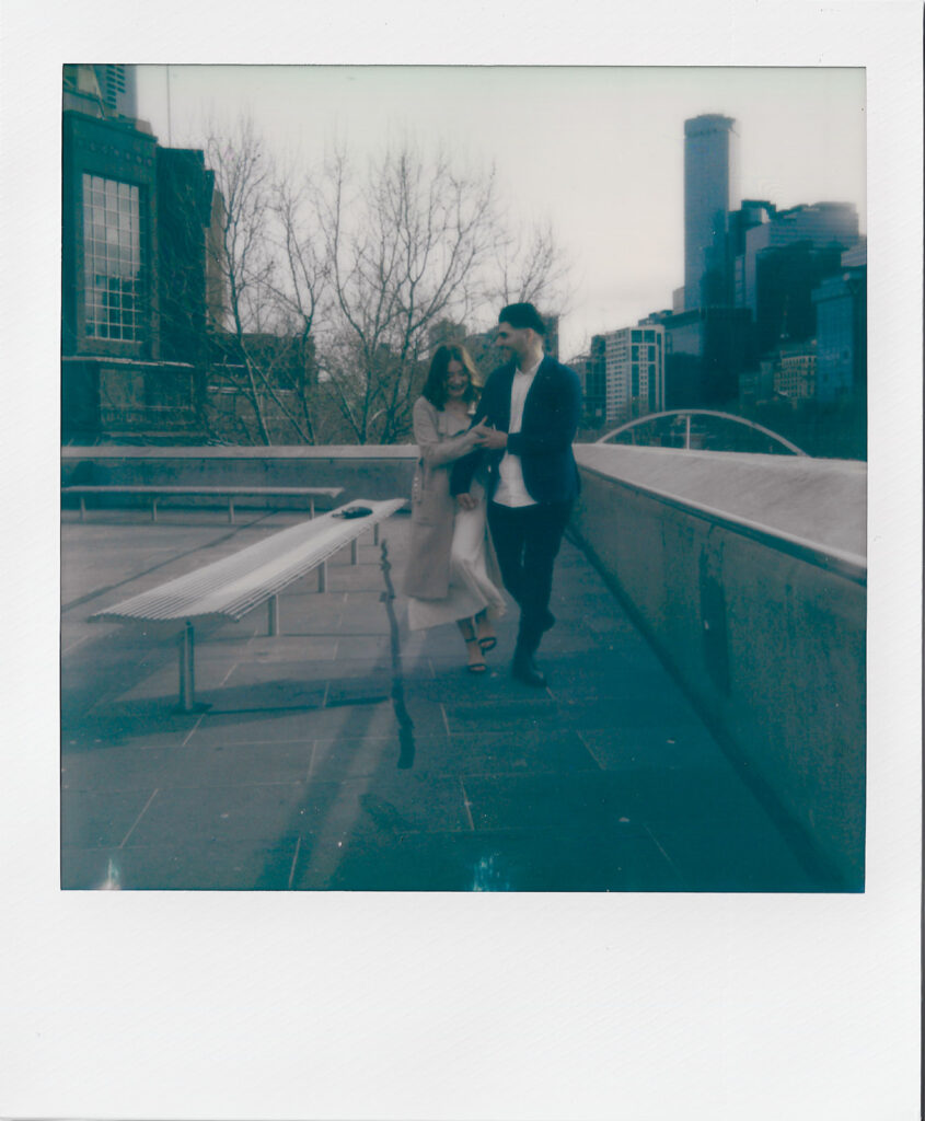 a polaroid photo of a couple holding hands during their pre-wedding photos, walking and laughing together with the southbank melbourne cityscape behind them