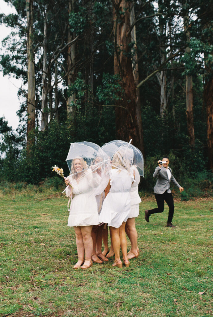 a film photo of a group of bridesmaids huddling together under clear umbrellas while a groomsman is filming them on a camcorder