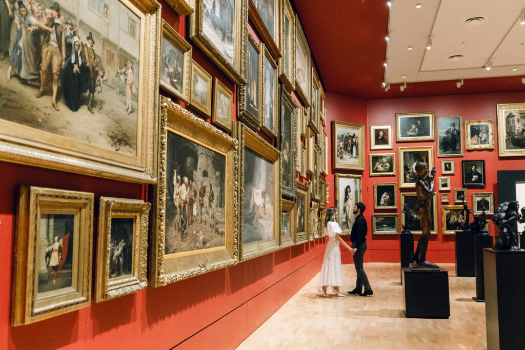 a red room with classic paintings hanging in goldframes, a couple is visible in the background holding hands