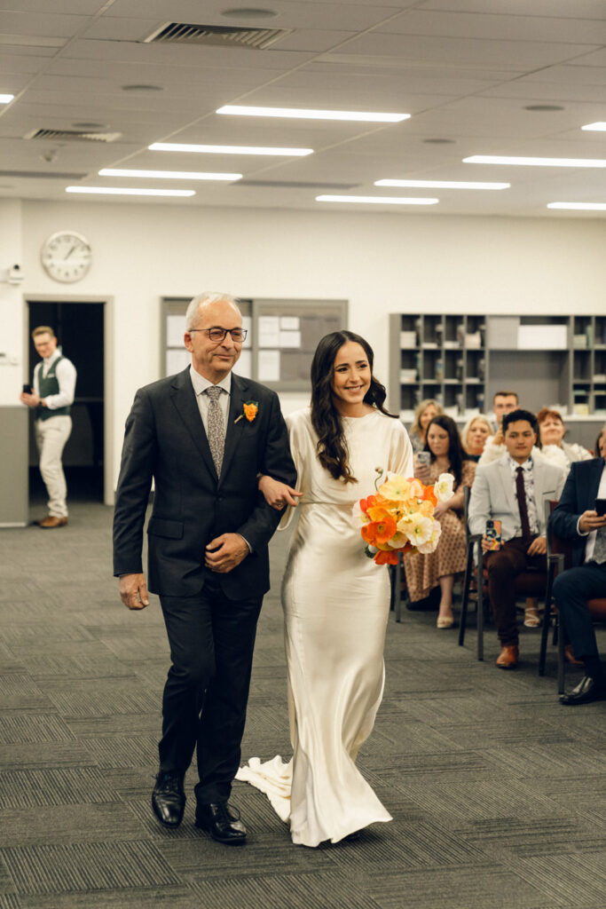 Bride walking down the aisle with a silk log sleeve gown and an orange poppy bouquet