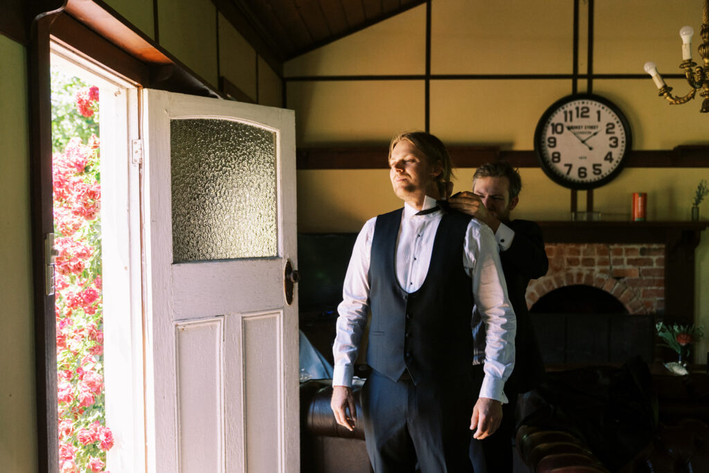 a groomsman helping the groom put on his bow tie in the rumpus room of ravenswood homestead